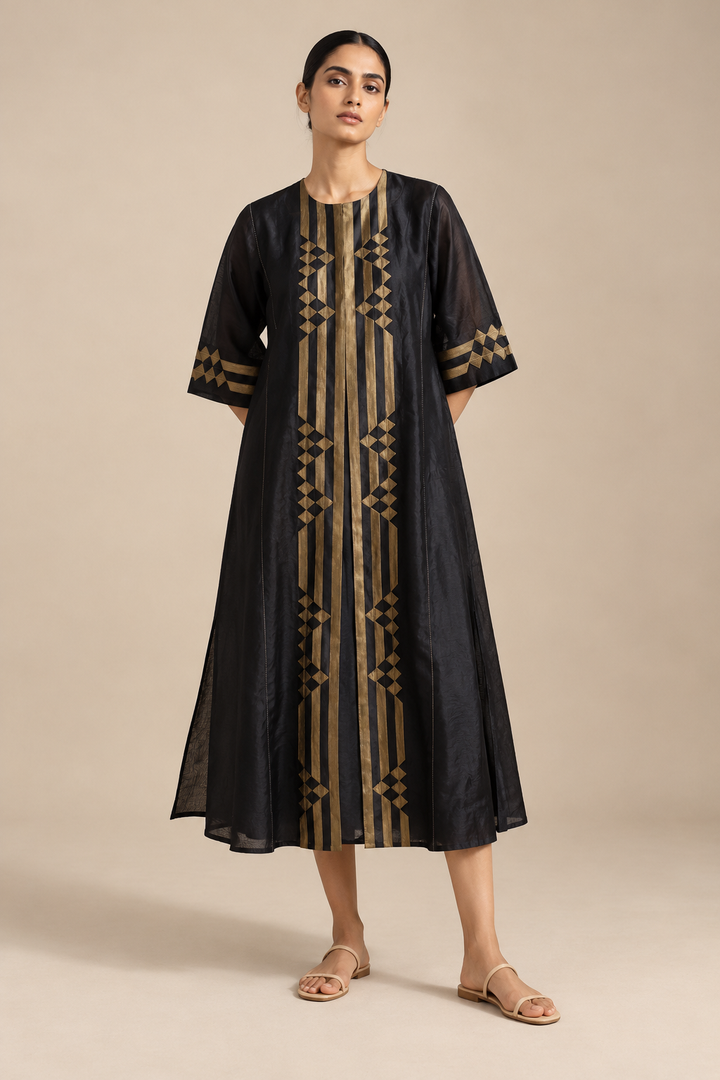 A woman modeling a black midi kurta with vertical gold stripes and geometric patterns on the front, along with matching patterned sleeve cuffs, set against a solid beige studio background.