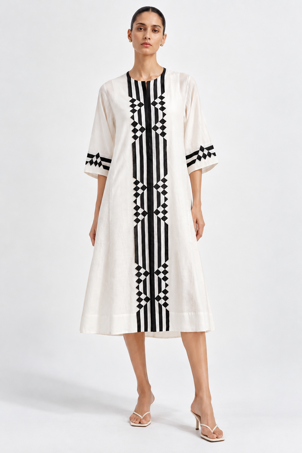 A woman modeling a cream-colored midi dress with bold black vertical stripes and geometric diamond patterns on the front, sleeves, and neckline, paired with neutral heeled sandals against a light gray studio background.
