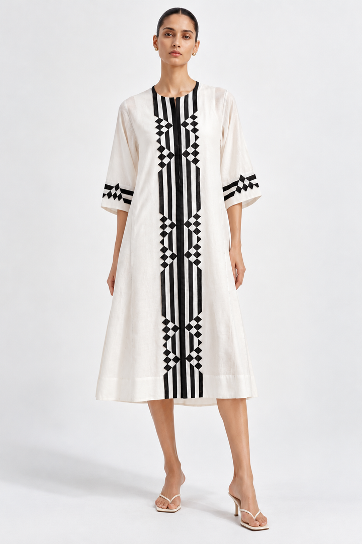 A woman modeling a cream-colored midi dress with bold black vertical stripes and geometric diamond patterns on the front, sleeves, and neckline, paired with neutral heeled sandals against a light gray studio background.