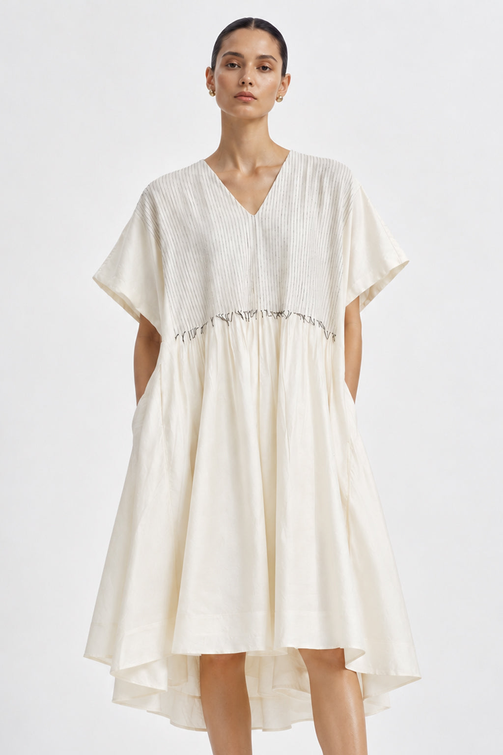 A woman modeling a cream-colored, high-low tiered dress with short sleeves and a gathered waist, set against a plain light gray studio background.